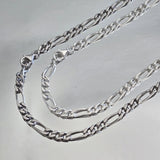 Comparaison visuelle entre chaîne homme argent massif rhodié (plus sombre) et argent massif blanc (traditionnel) maille Figaro 3 mm alternée.